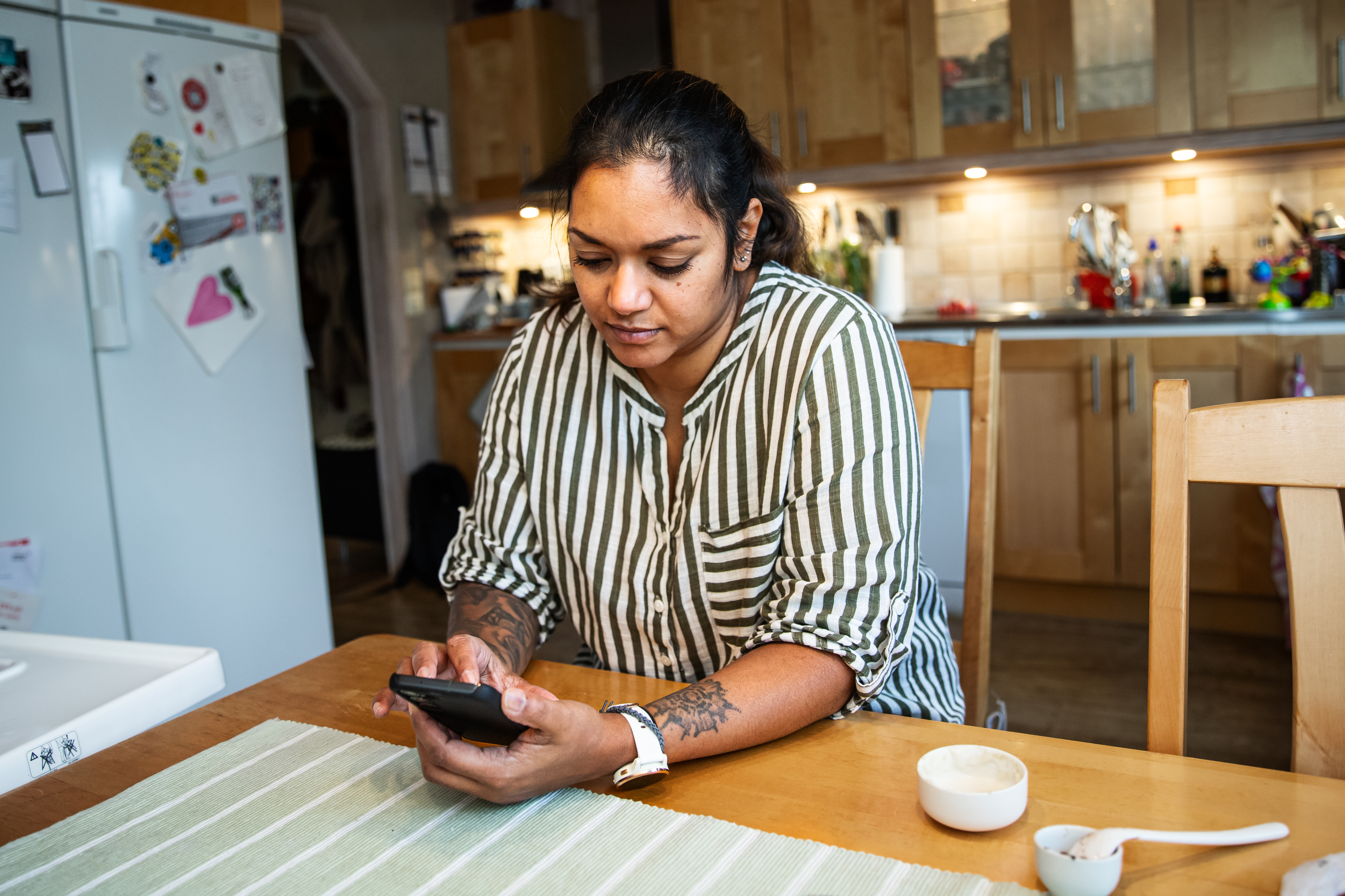 Kvinna i randig skjorta sitter vid köksbord och tittar i mobil.
