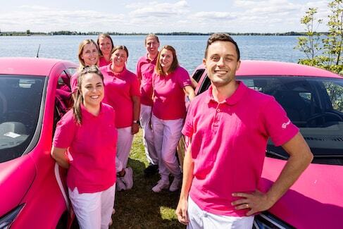 Sju medarbetare på Hälsa Hemma står framför en sjö och har på sig rosa pikétröjor och vita byxor. De står mellan två rosa bilar.