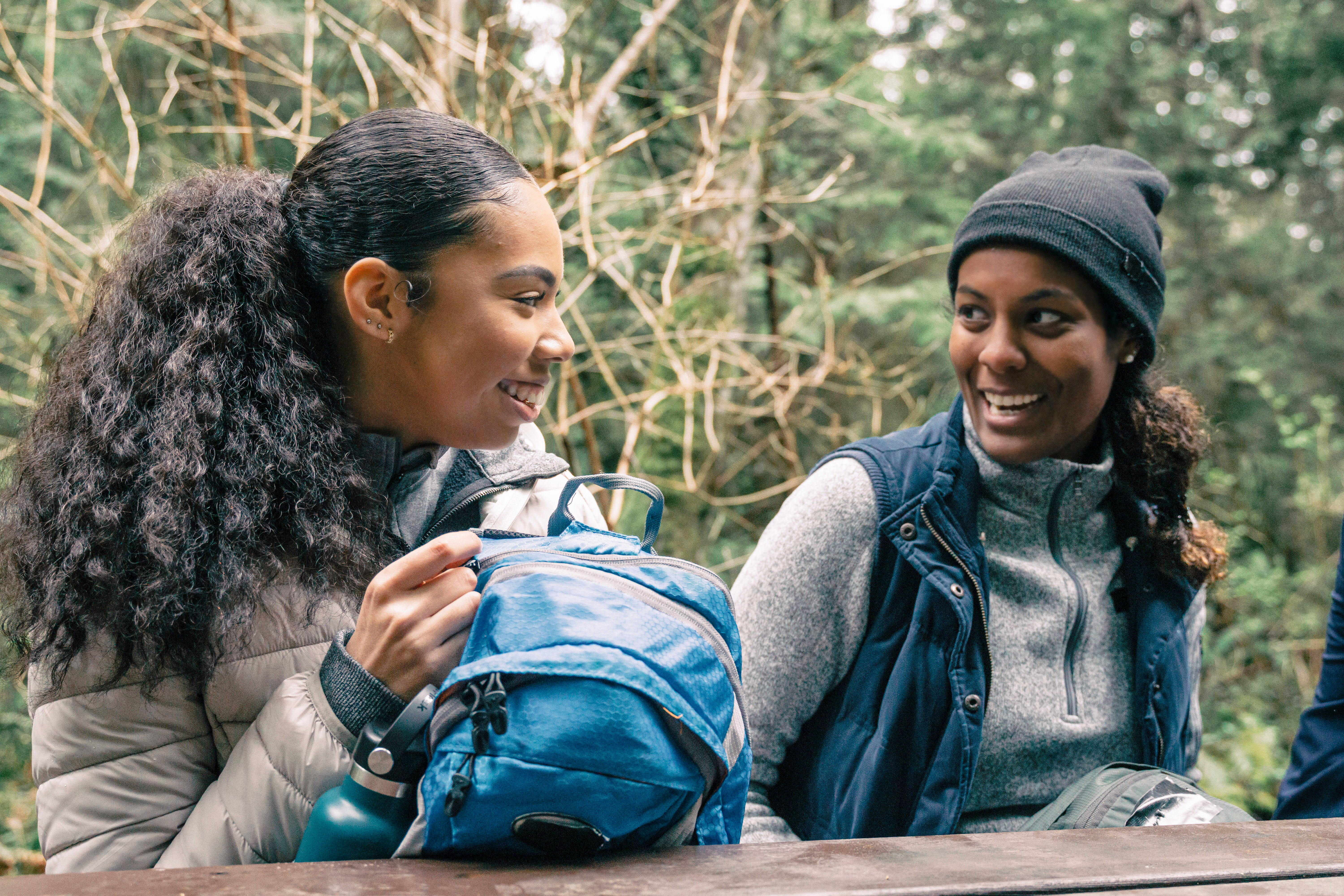 Två tjejer sitter vid ett bord i skogen och ler mot varandra.