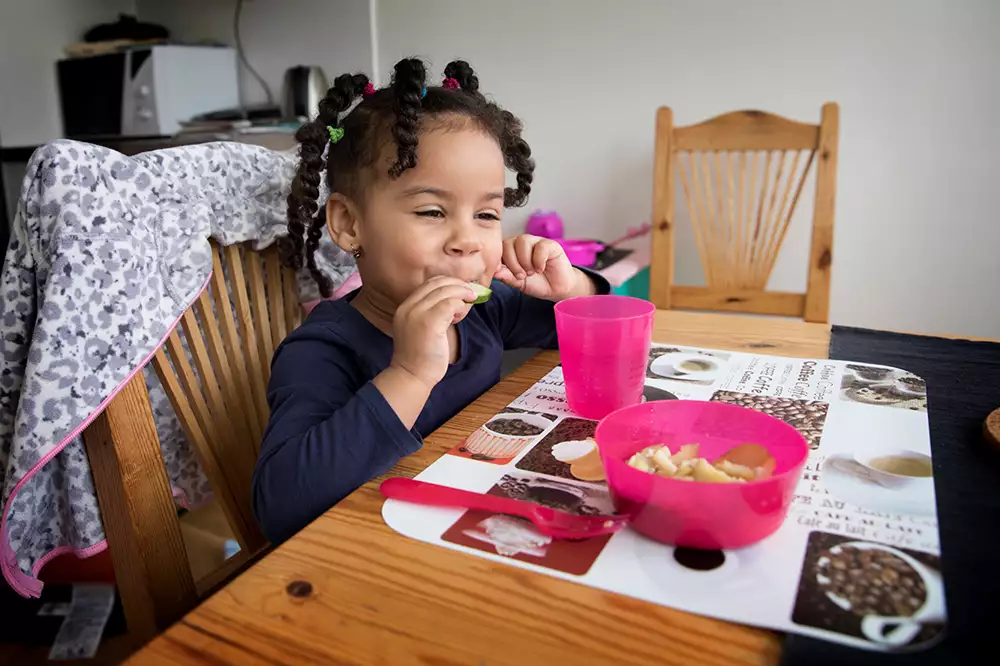 Barn som äter en fruktbit och har en skål med fruktbitar på köksbordet