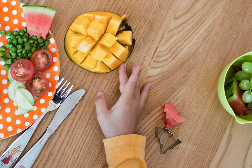 Ett bord med uppskuren frukt och grönsaker. Ett barns hand petar på en mango.