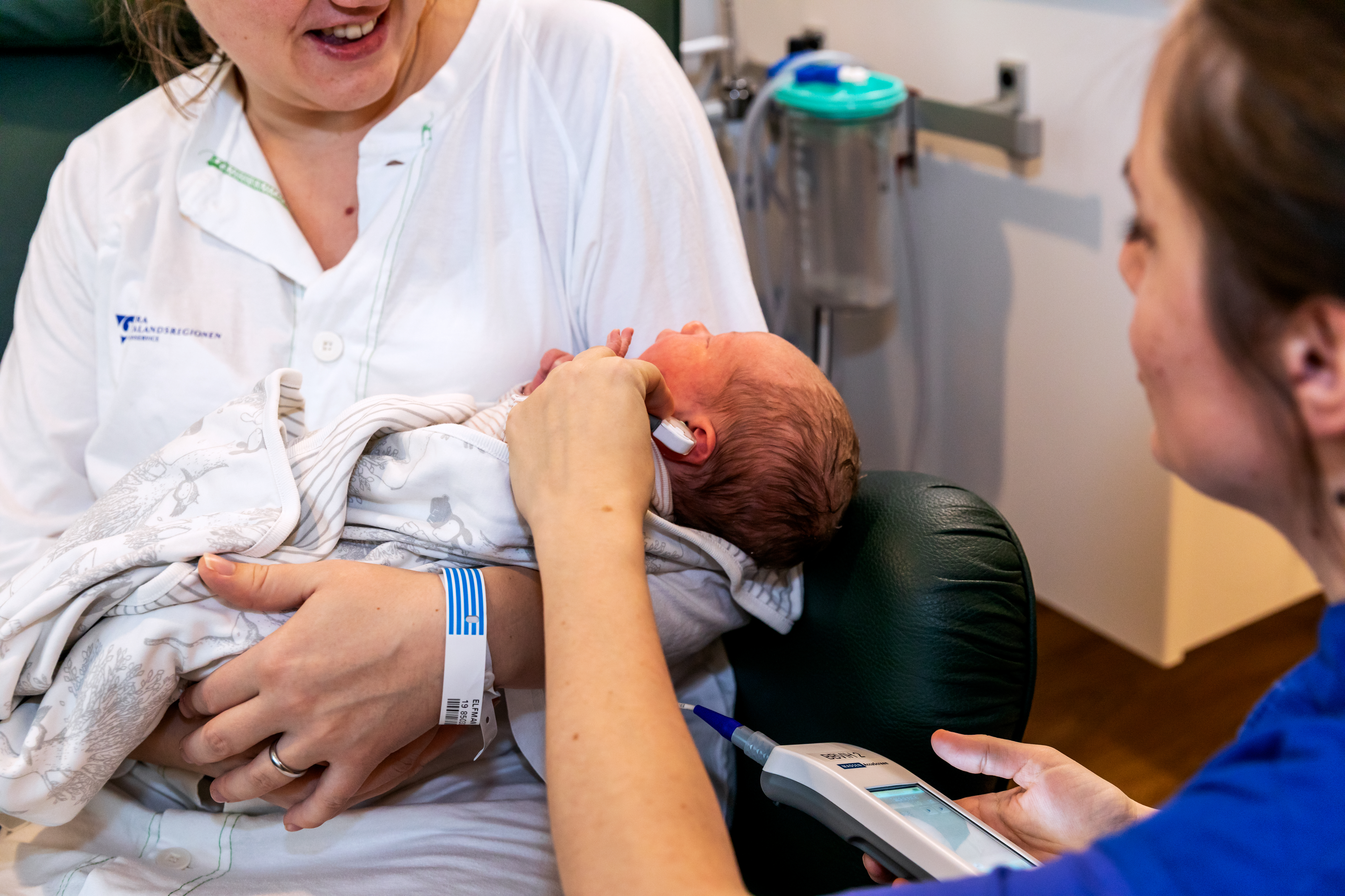 Ett nyfött barn ligger i famnen på en vuxen, en barnmorska mäter i barnets öra.