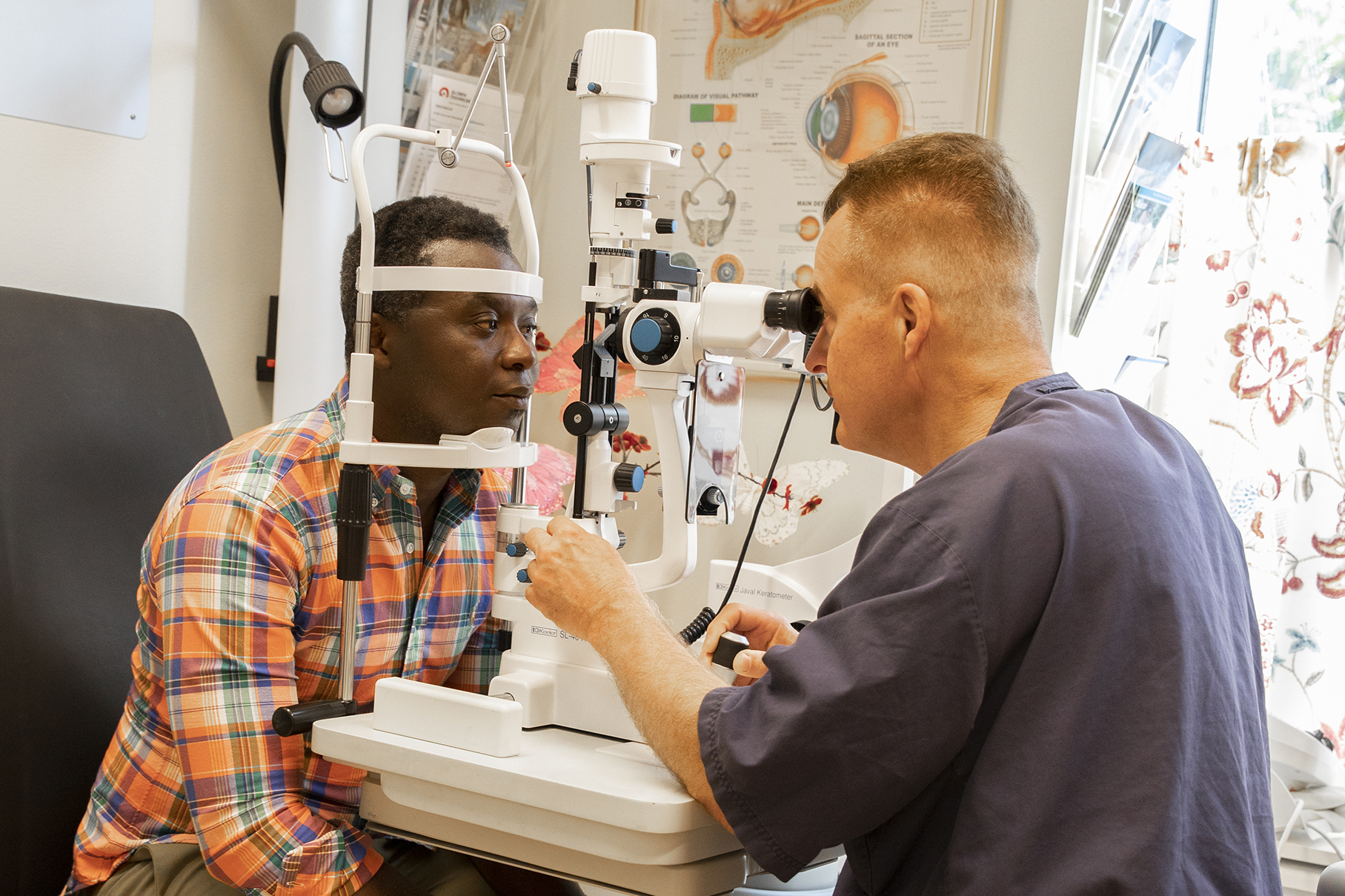 En person sitter och tittar in i ett ögonmikroskop och läkaren sitter på andra sidan.