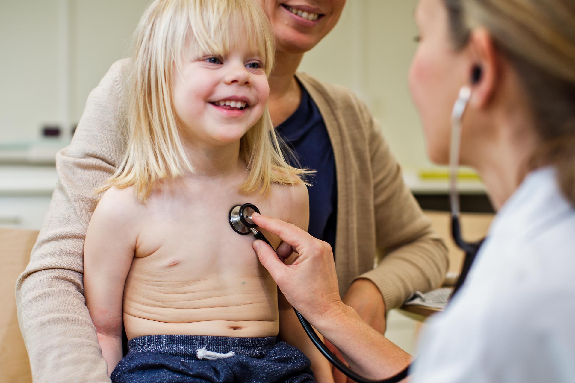 Barnläkare lyssnar på hjärtat med stetoskop på ett barn som sitter i knät på en förälder.
