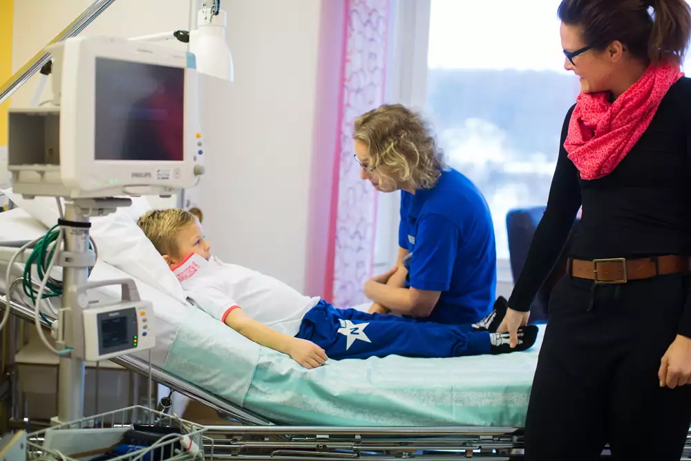 Ett barn ligger i en sjukhussäng. En vårdperson sitter bredvid på sängen och en förälder står vid sängen och håller barnet i foten.