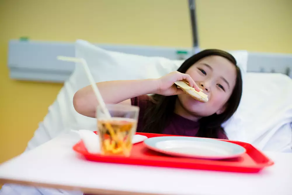 Barn som sitter i en sjukhussäng med en matbricka och äter en smörgås.
