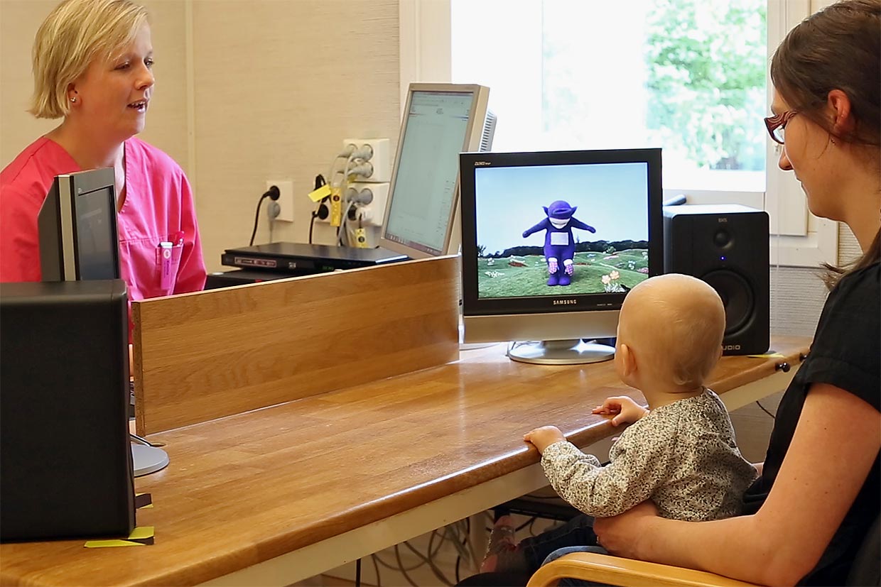Ett litet barn tittar på en bildskärm med en intressant lila figur under en hörselundersökning. En audionom och barnets förälder finns också i rummet.