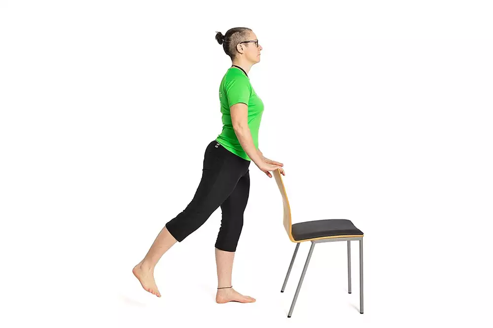 En person står bakom en stol och sträcker ena benet snett bakåt.