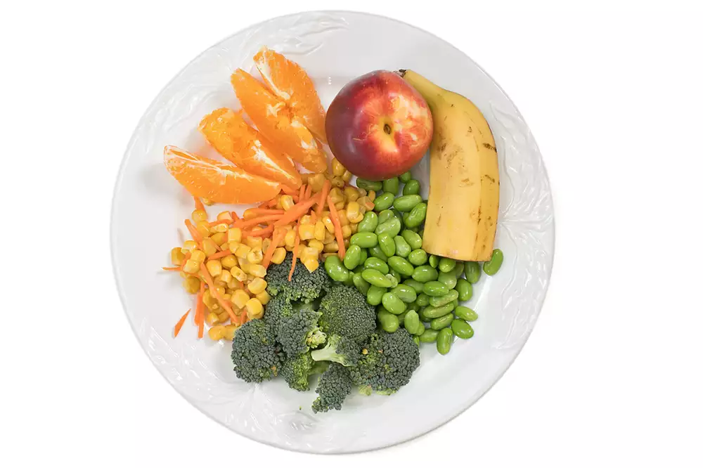 En tallrik med olika frukter och grönsaker, bland annat apelsin, banan, sojabönor, broccoli och majs.