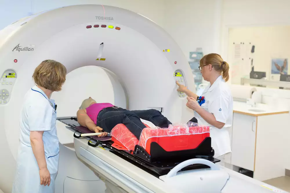 Patient som ligger på en brits som förs in i en strålbehandlingsapparat. Två vårdpersoner står vid sidan av, varav en trycker på en knapp.