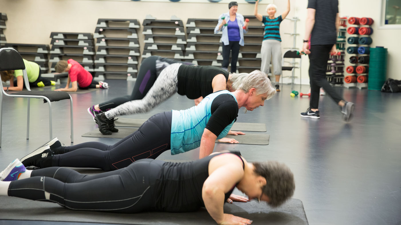 En grupp tränar i en träningslokal. De ligger på magen med mattor under sig på golvet och gör en rörelse.