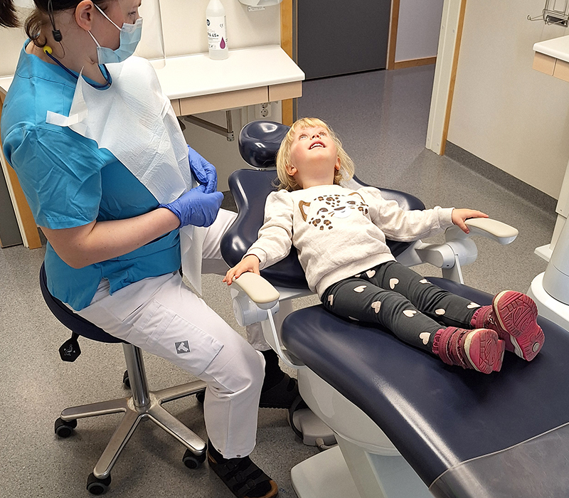 Liten flicka ligger ner i tandläkarstol och tandläkare sitter bredvid.