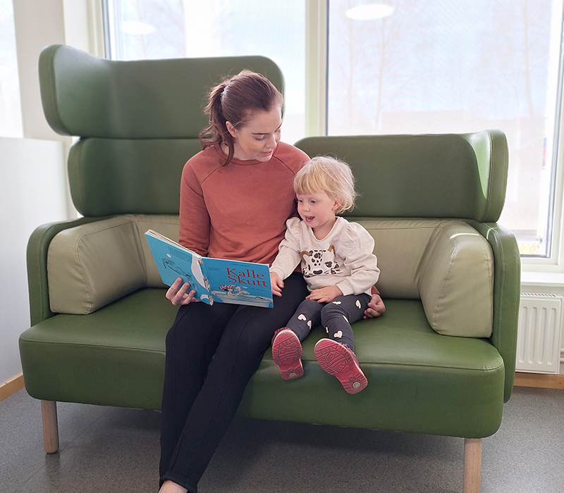 Mamma och dotter sitter i en grön soffa i väntrummet och läser bok.