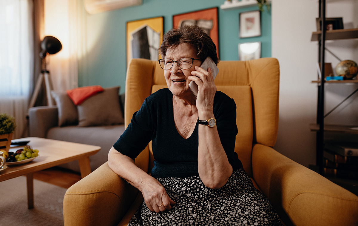 Äldre kvinna som pratar i telefonen med ett leende på läpparna. Sitter i fåtölj.