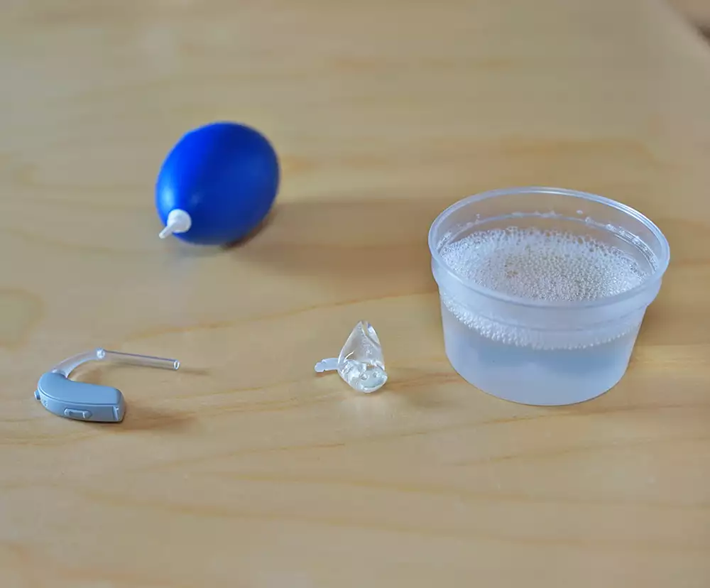 En hörapparat med gjuten insats, ett glas med diskmedel och vatten i, och en kanylblåsa på ett bord.