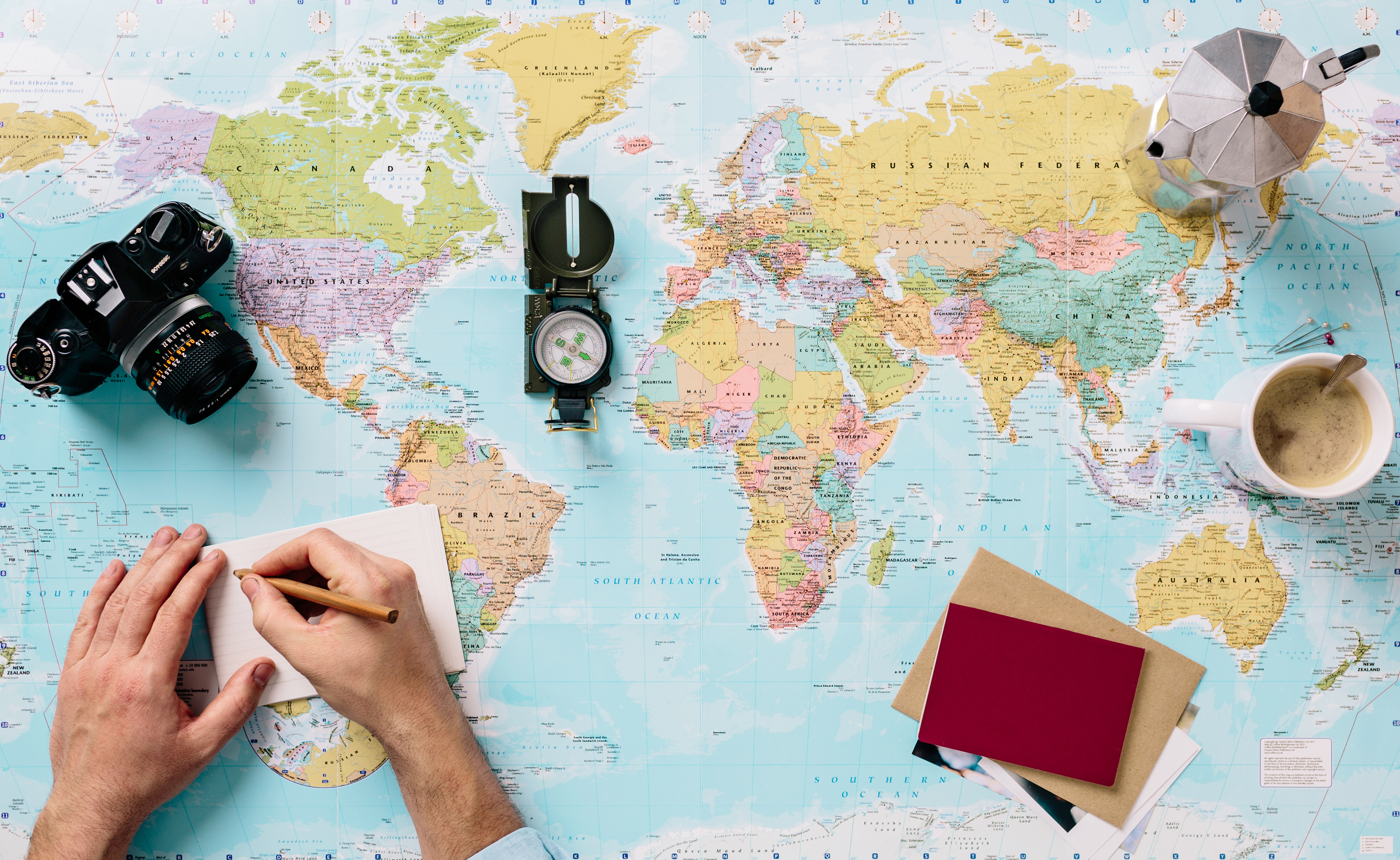 Bild på en världskarta och en persons händer som planerar resa. Med på bilden finns även en kaffekopp, pass och kamera.