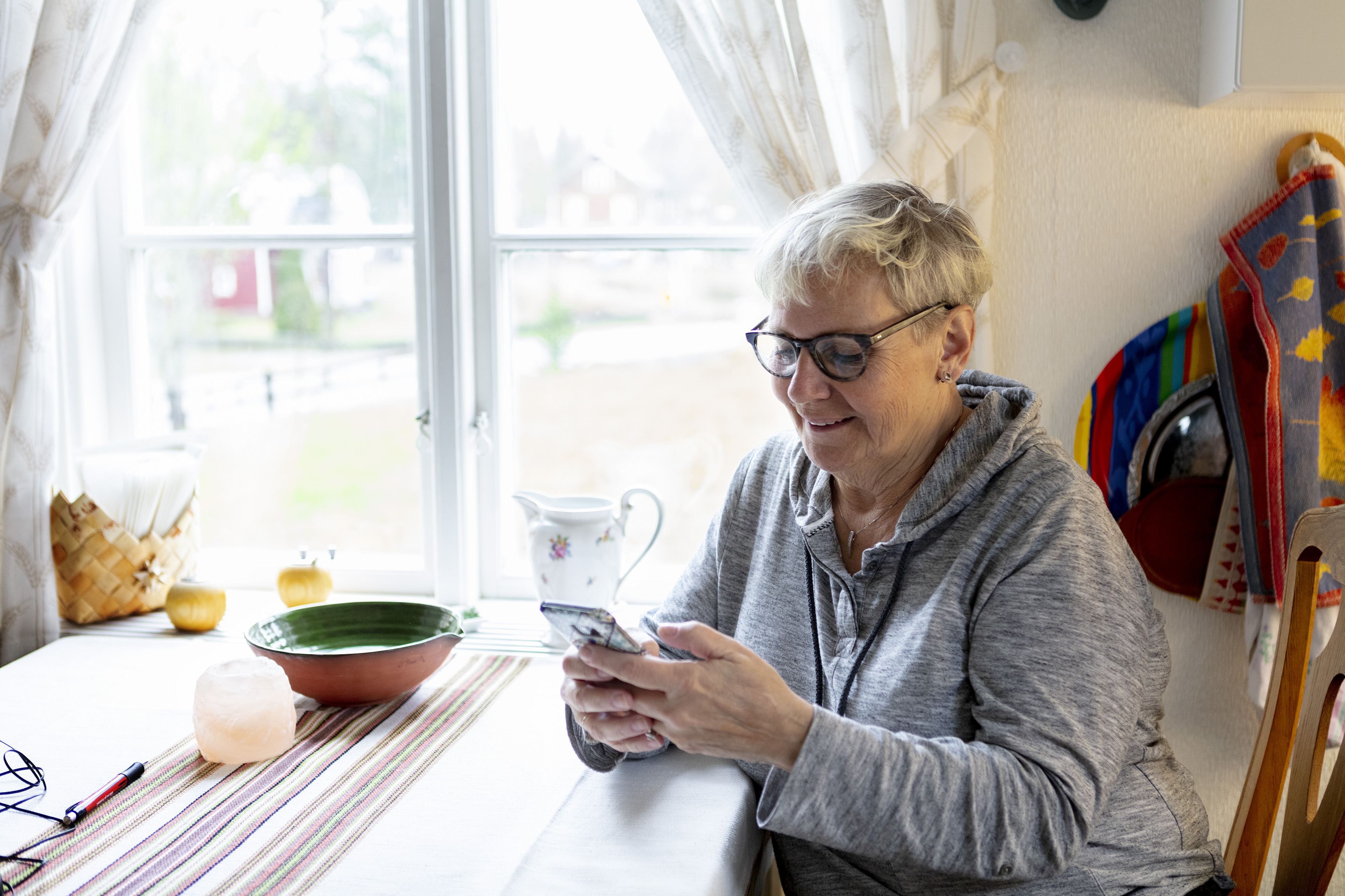 Dam med mobil i handen sittande vid sitt köksbord hemma