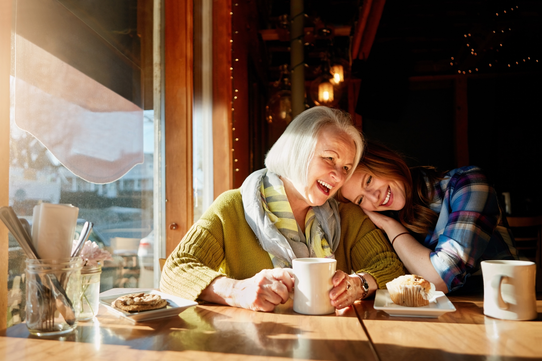 Äldre kvinna och yngre kvinna tillsammans vid ett bord där yngre kvinna lutar sig mot den äldre.