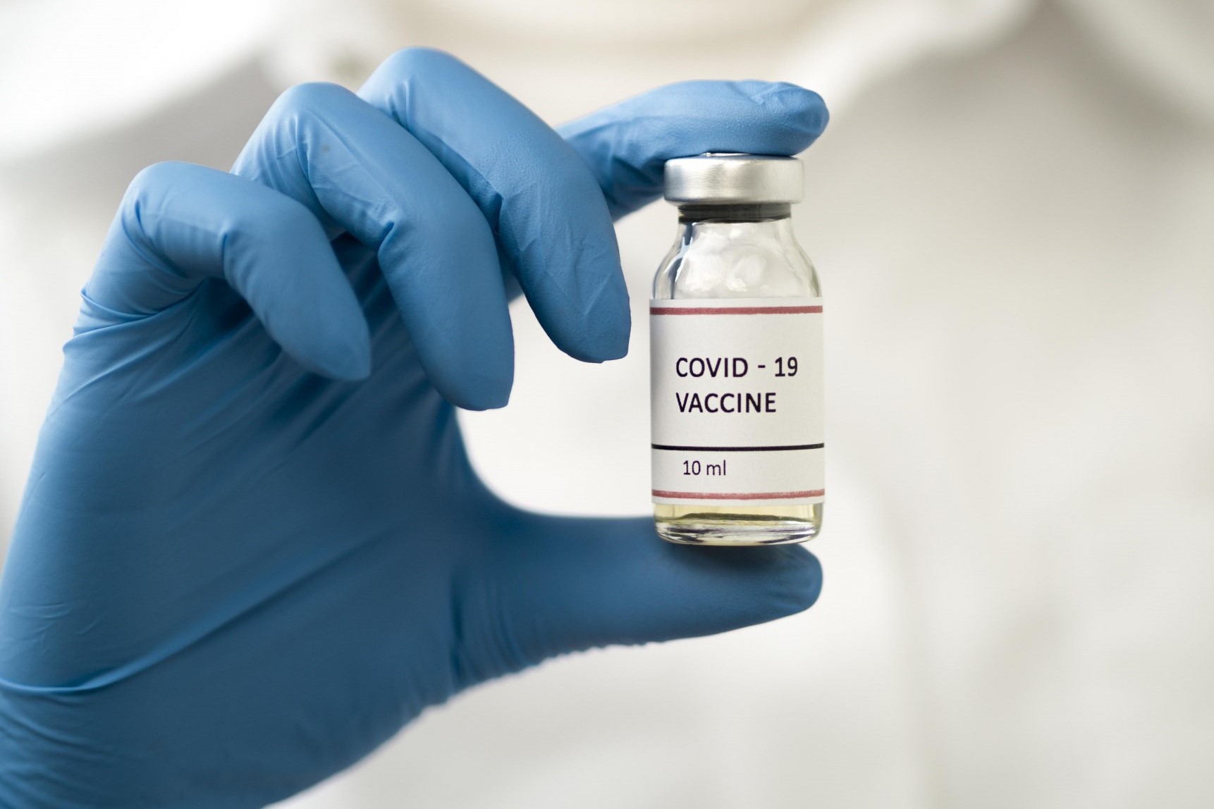 Person håller fram en vaccinflaska med text covid-19 vaccine, har blå plasthandske och vita kläder