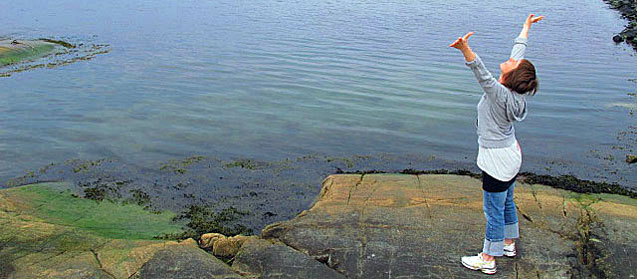 Dansterapeut Marit Mörk står på en klippa vid vattnet och sträcker upp händerna
