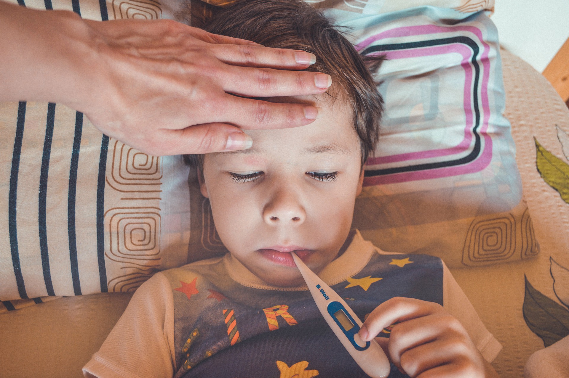 Sjukt barn med febertermometer i munnen. Vuxen person lägger sin hand över barnets panna.
